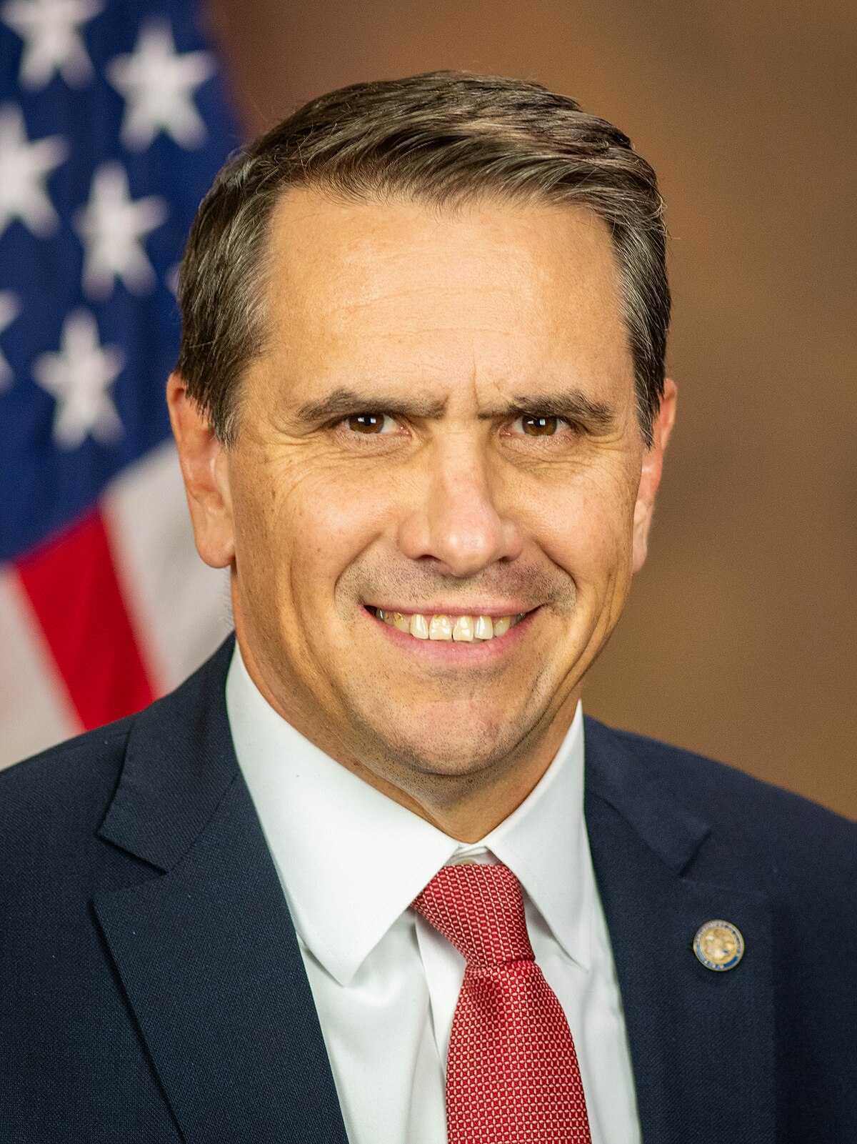 Official portrait of Todd Blanche in a dark suit and red tie before a blurred United States flag.