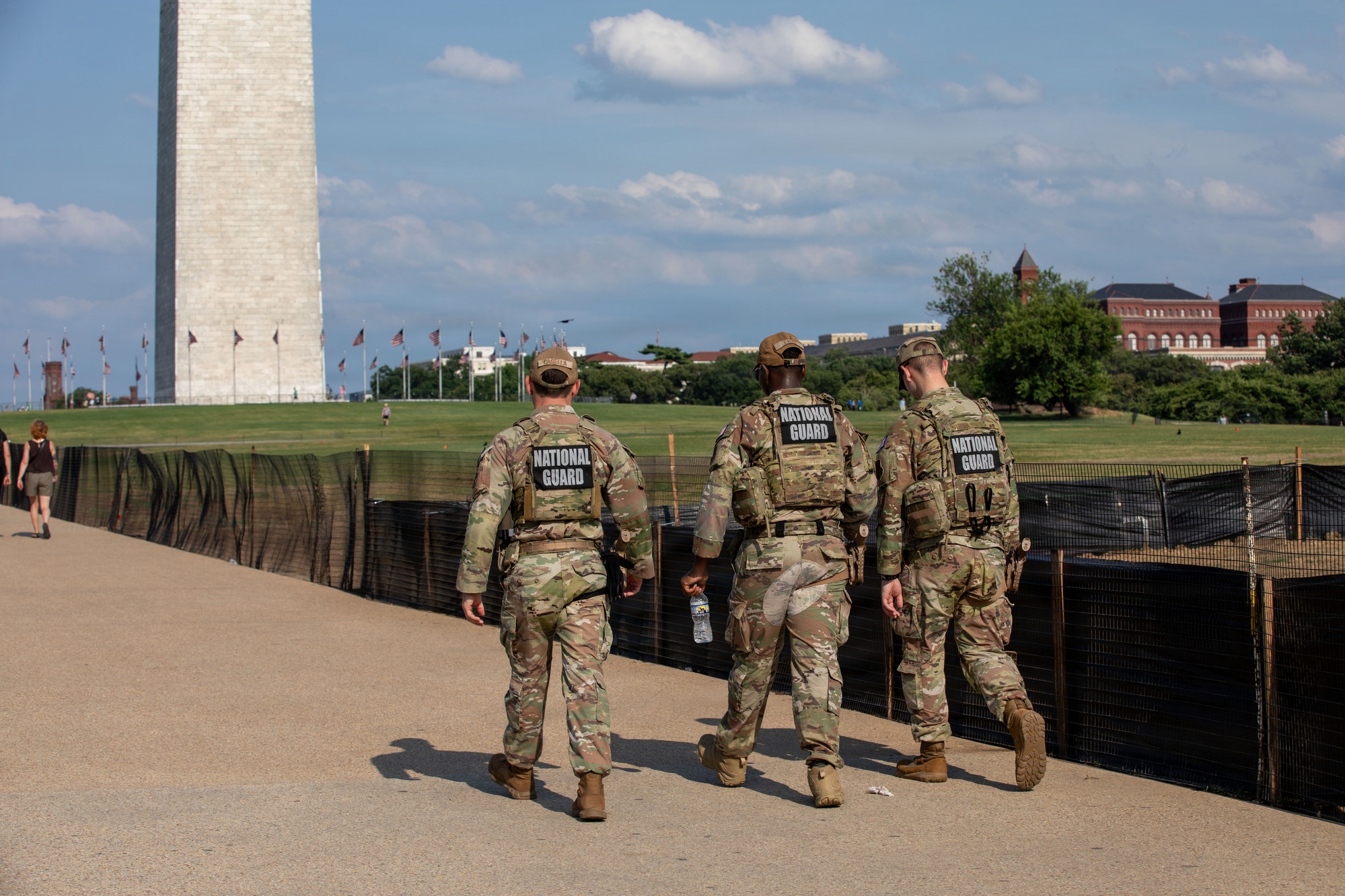 Three National Guard members walk on the National Mall with the Washington Monument at left and the Smithsonian Castle in the distance.